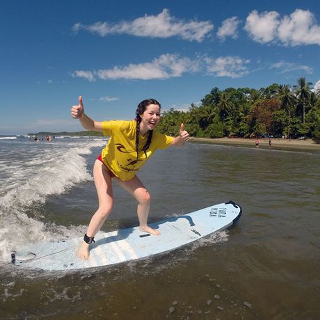 Surfen in Ballena EIne junge Frau steht mit Daumen hoch auf dem Surfbrett und lässt sich von einer Welle in Richtung Strand tragen.